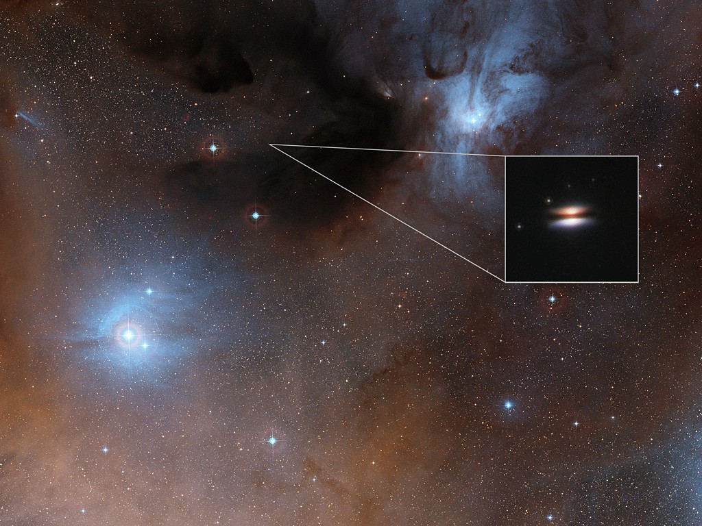 The young star 2MASS J16281370-2431391 lies in the spectacular Rho Ophiuchi star formation region, about 400 light-years from Earth. It is surrounded by a disc of gas and dust — such discs are called protoplanetary discs as they are the early stages in the creation of planetary systems. This particular disc is seen nearly edge-on, and its appearance in visible light pictures has led to its being nicknamed the Flying Saucer. The main image shows part of the Rho Ophiuchi region and a much enlarged close-up infrared view of the Flying Saucer from the NASA/ESA Hubble Space Telescope is shown as an insert.